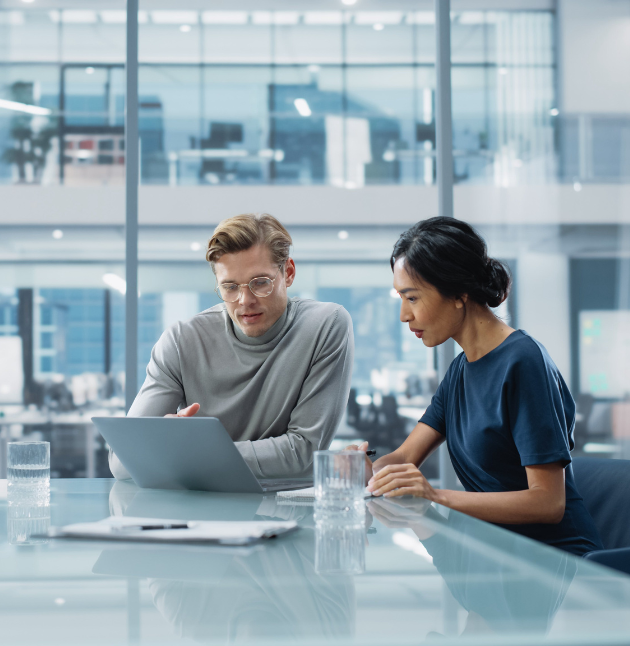 Two people sit at a glass table in an office, focused on a laptop for tax planning. Glasses and a pen are on the table. Office windows and a modern interior are in the background.